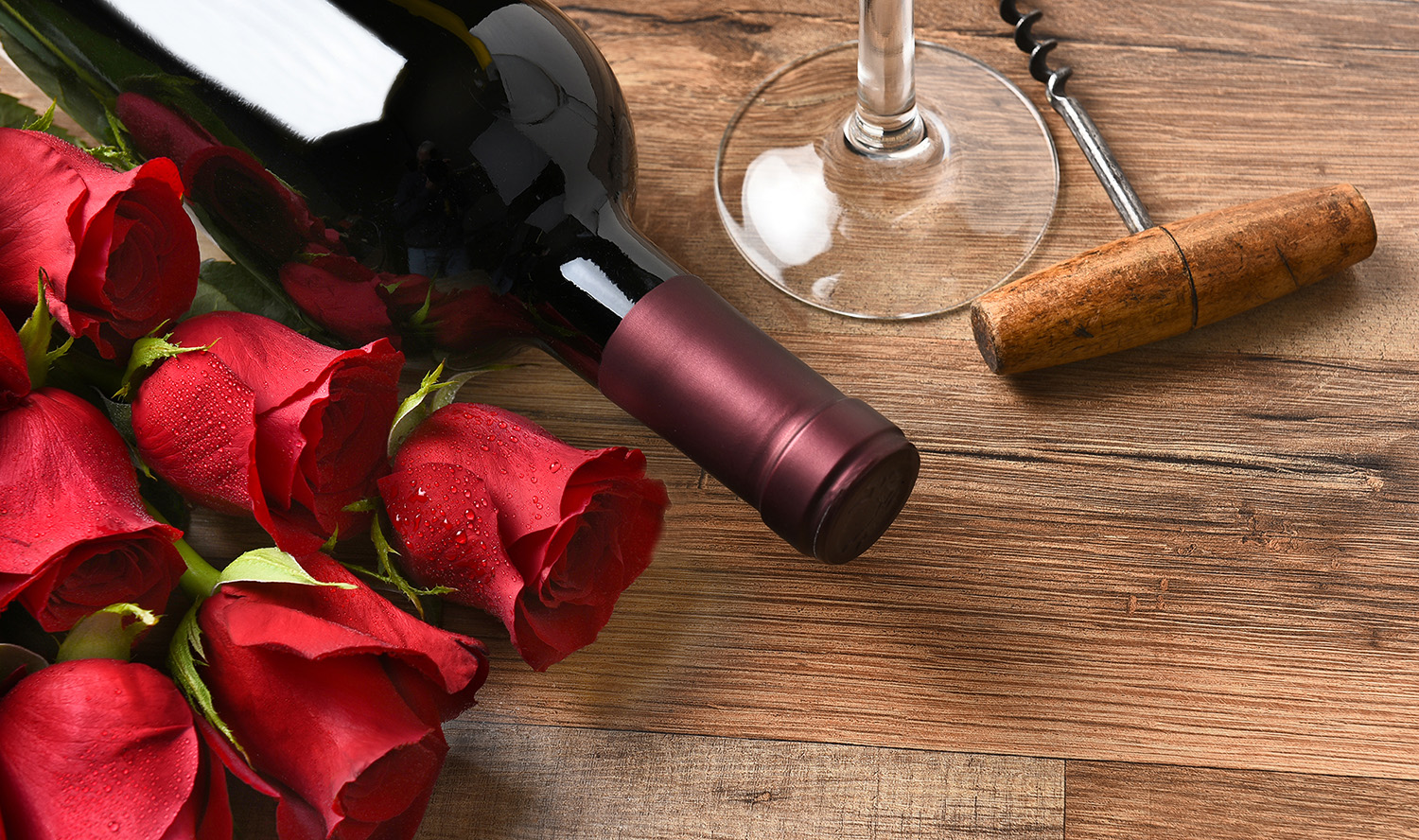 A wine bottle with red roses and a glass and cork screw on a rustic wood table.